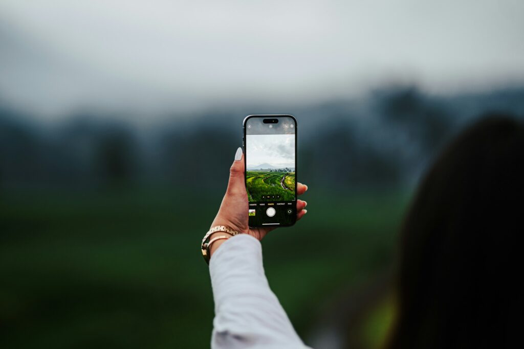 Hand hält Smartphone und fotografiert eine Wiese vor bewaldeten Hügeln